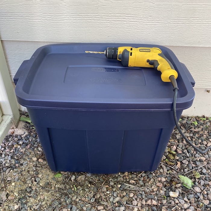 A large Plastic bin with a drill machine is placed on rocks.