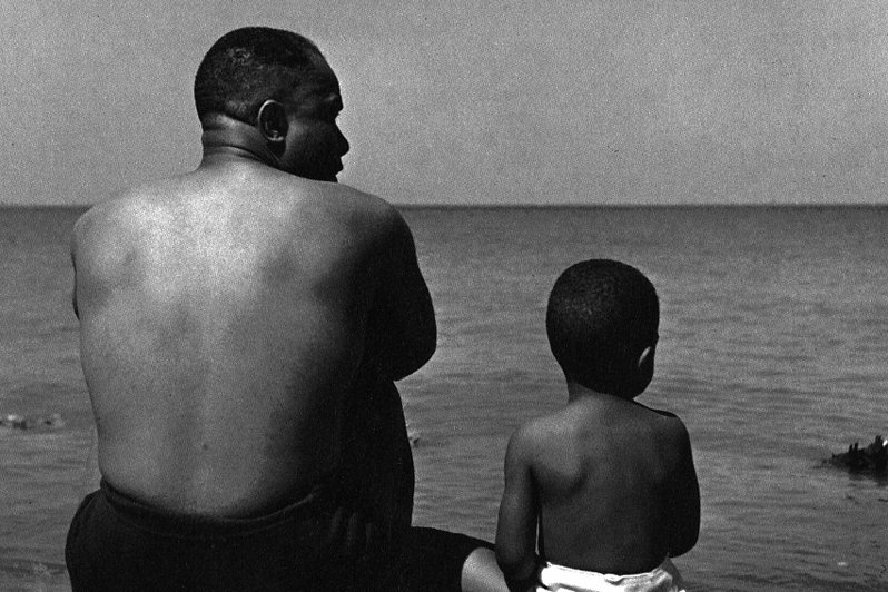 Shirtless man and a kid sitting in front of ocean.