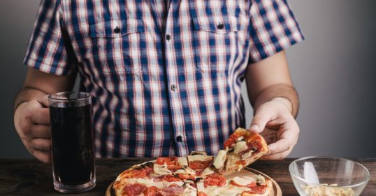 A man is holding a slice of pizza and a glass of beer, wondering what to do about overeating.
