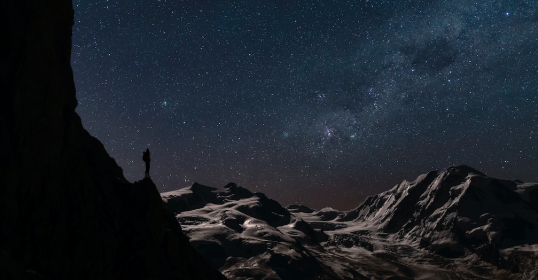 A person standing on top of a mountain with the milky way in the sky, contemplating existence.