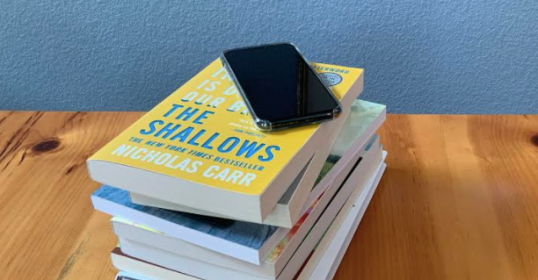 A stack of books with a cell phone on top, showcasing the intersection of traditional books and digital life.