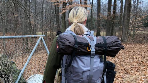 A person with blonde hair is seen from behind, wearing a backpack with a rolled-up sleeping bag attached, walking through a wooded area next to a chain-link fence.