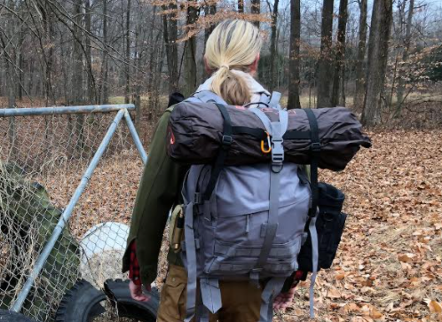 A person with blonde hair is seen from behind, wearing a backpack with a rolled-up sleeping bag attached, walking through a wooded area next to a chain-link fence.