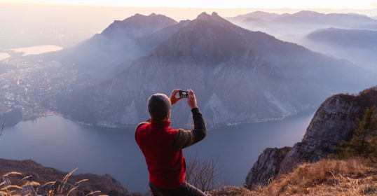 A man using his smartphone to take a photo of a mountain and lake.