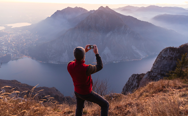 A man using his smartphone to take a photo of a mountain and lake.