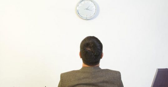 A man at a desk, gazing at a clock, is a prime example of procrastination and its effects on behavior.