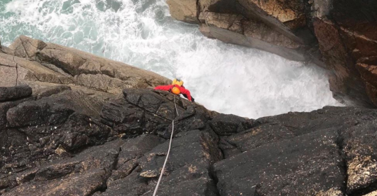 A man is climbing up a rocky cliff near the ocean, feeling uncomfortable yet determined.
