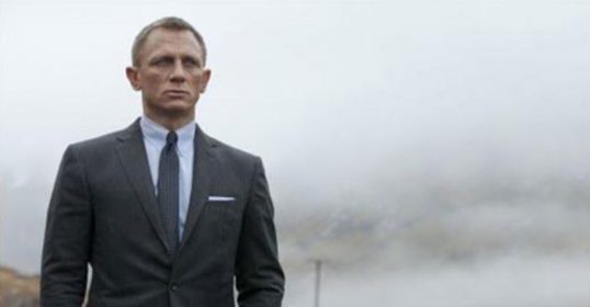 James Bond, displaying stoicism, stands next to a car in a suit.