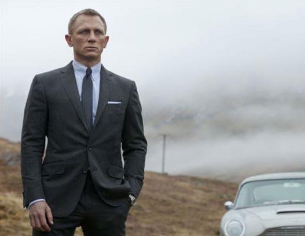 James Bond, displaying stoicism, stands next to a car in a suit.