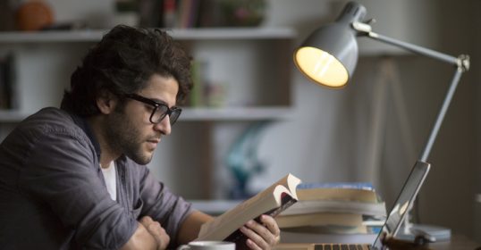 A man is immersed in a book at a desk with his laptop.