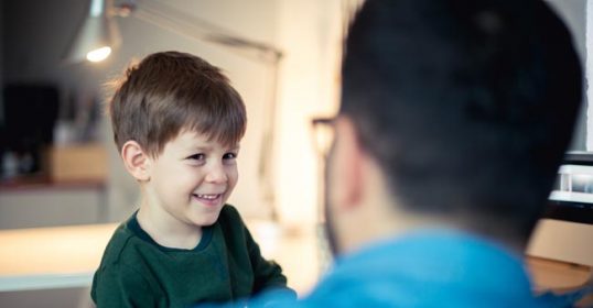 A little boy is happily smiling at a man in front of a computer.