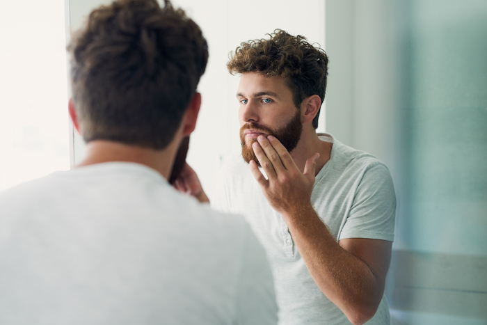 A man is brushing his beard in front of the mirror while pressing his beard oil.
