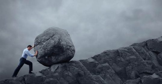A businessman struggling to push a large rock up a mountain.