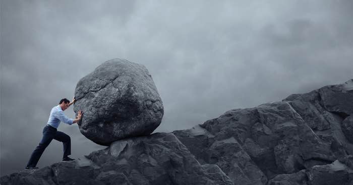 A businessman struggling to push a large rock up a mountain.