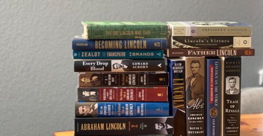 A stack of biography books on a wooden table.