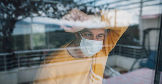 A man wearing a medical mask looks out of a window during a crisis.