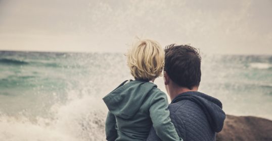 A man is holding a child while gazing at the ocean.