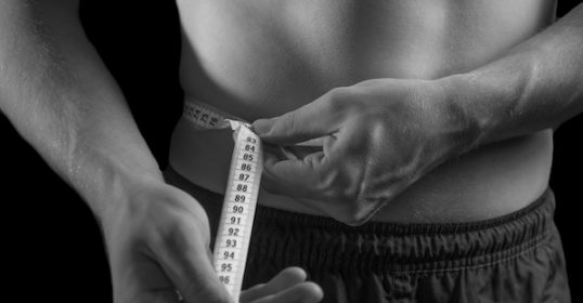 A man is measuring his waist with a measuring tape to track his body fat.
