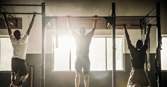 A group of men doing pull ups in a gym to reach their physical benchmarks.