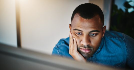 A man, bored, staring at a computer screen with his hand on his face.