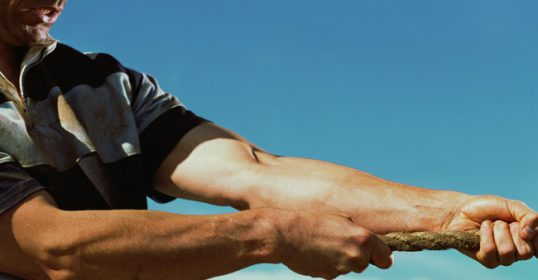 Close-up of two people engaged in a tug of war, highlighting their arms and the rope as they develop grit.