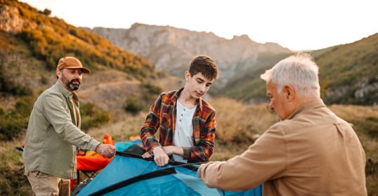 A father is setting up a tent in the mountains, initiating his family into the beauty of nature.