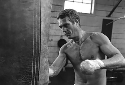 A black and white photo of a man punching a workout bag, reminiscent of Steve McQueen.