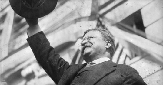 A man in a suit and hat, channeling the Last Romantic, is holding up a hat.