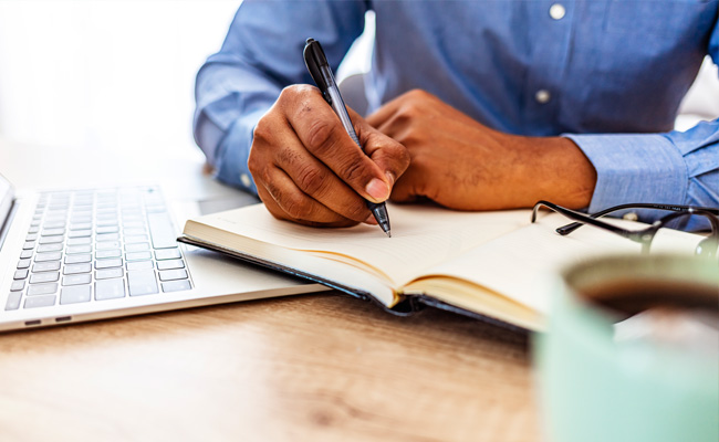 A man's hand writing in a notebook next to a laptop.