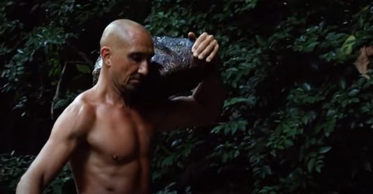 A shirtless man, older and rugged, carrying a large rock in the jungle.