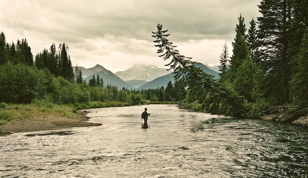 A man is fly fishing in a river with mountains in the background, reflecting on life lessons.