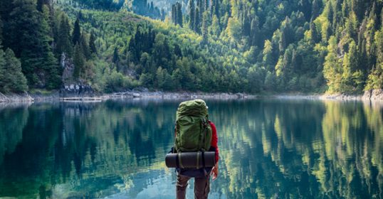 A man with a backpack is standing on a rock near a lake, enjoying the serene view.