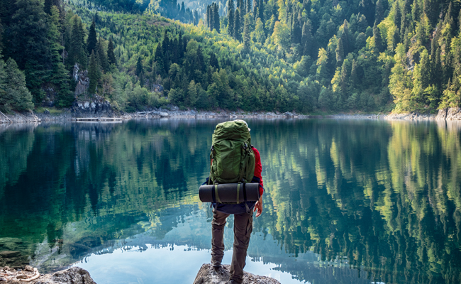 A man with a backpack is standing on a rock near a lake, enjoying the serene view.