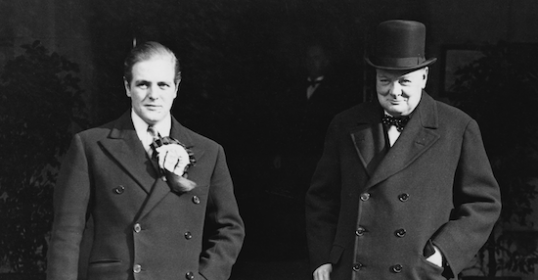 Two men in suits standing next to each other, possibly discussing a podcast or sharing a relationship.