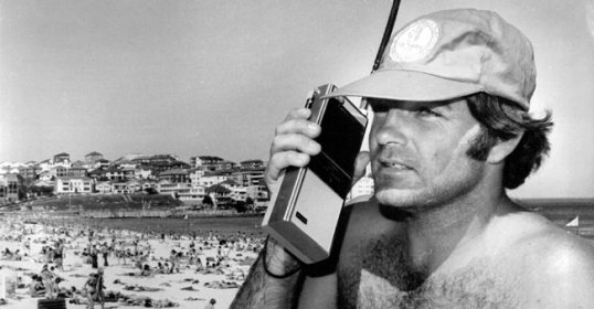 An introduction of a man in a hat using a walkie talkie on the beach.