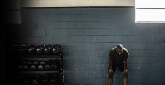 A sore man leaning against a wall in a gym, wondering what to do after his workout.