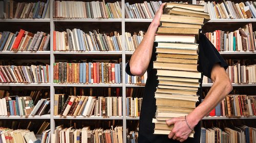 A man holding a stack of books in front of his home library.