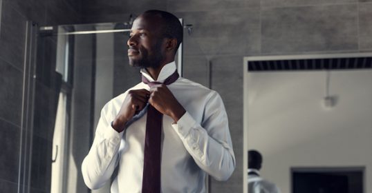 A man is adjusting his necktie in a bathroom.