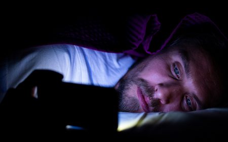 A man laying in bed with a cell phone, browsing the internet.