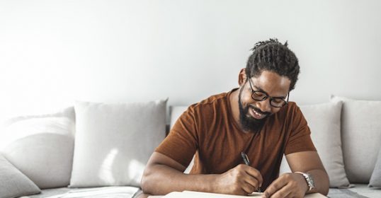 A black man writing in a notebook at home.