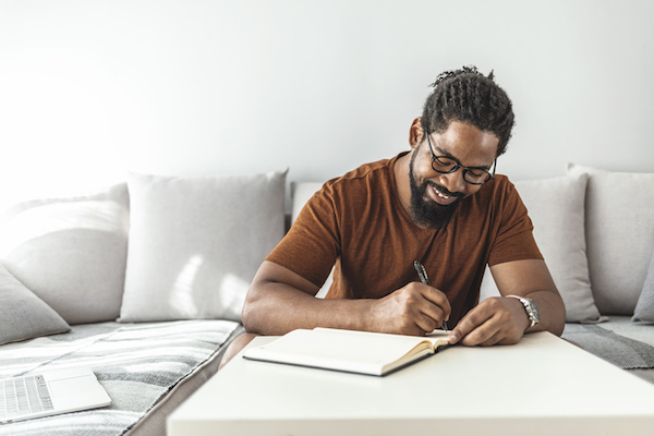 A black man writing in a notebook at home.