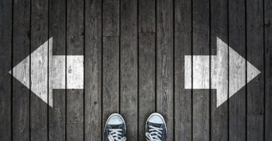 A person standing on a wooden floor with two arrows pointing in different directions, trying to make a big decision.