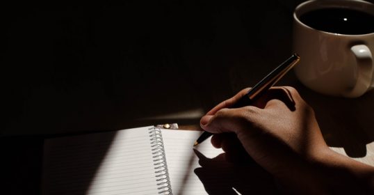 A hand writing keywords in a journal next to a cup of coffee.