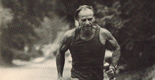 A man from the 1980s is jogging along a road with dumbbells, working on his walking workout to get in shape.