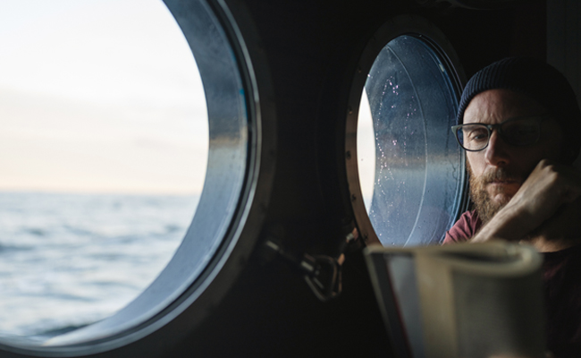 A bearded man gazes out the window of a ship.