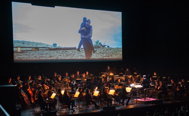 A stage with an orchestra and a screen showing a film score.