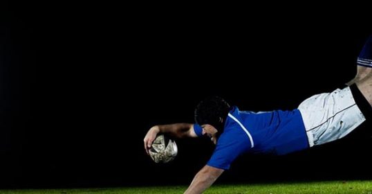 A rugby player is diving for the ball at night with tremendous effort.
