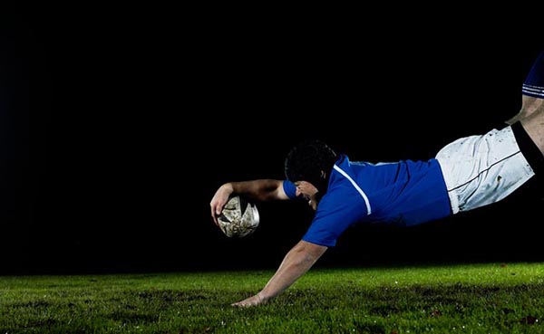 A rugby player is diving for the ball at night with tremendous effort.