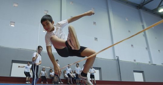 A boy is jumping over a high bar in a gym during P.E. class.