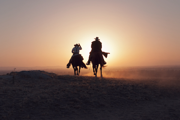 Two cowboys kicking up dust on their horses at sunset.